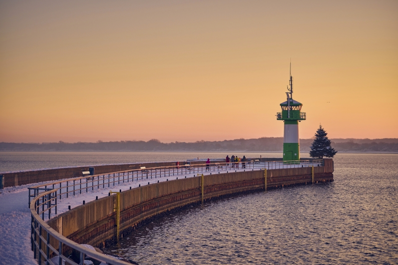 Eine weitläufige, schneebedeckte Mole führt in einem sanften Bogen zum grün-weißen Leuchtturm (Molenfeuer) von Travemünde. Direkt neben dem Turm steht ein geschmückter Weihnachtsbaum am Ende der Hafenmauer. Der Himmel ist in die warmen, orangefarbenen Töne eines winterlichen Sonnenuntergangs getaucht, während das ruhige Wasser der Ostsee den Kai umgibt.