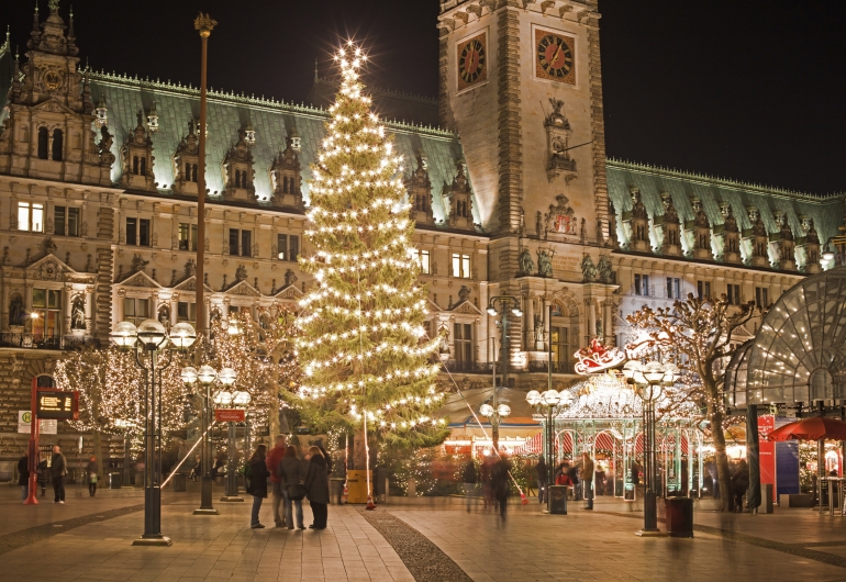 Eine Nachtaufnahme des Hamburger Rathausmarktes zur Weihnachtszeit. Im Bildzentrum steht ein hoher, hell erleuchteter Weihnachtsbaum mit einem leuchtenden Stern an der Spitze. Im Hintergrund ist die beleuchtete Fassade des Hamburger Rathauses mit seinem markanten Turm und der Uhr zu sehen. Rechts befinden sich Marktstände und ein dekorierter Pavillon mit einer leuchtenden Schlitten-Silhouette auf dem Dach. Passanten bewegen sich über den gepflasterten Platz, der von historischen Straßenlaternen beleuchtet wird.
