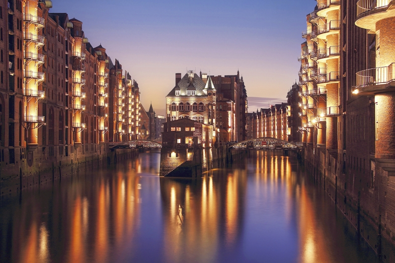 Eine abendliche Aufnahme der Hamburger Speicherstadt während der blauen Stunde. Im Zentrum steht das hell erleuchtete Wasserschlösschen auf einer Halbinsel, umgeben von einem Kanal (Fleet). Flankiert wird das Gebäude von hohen, mehrstöckigen Backsteinspeichern mit zahlreichen Balkonen, deren Fassaden warm beleuchtet sind. Die Lichter spiegeln sich deutlich im ruhigen Wasser des Kanals. Im Hintergrund sind zwei Brücken zu erkennen, die über das Fleet führen.
