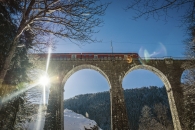 Eine Untersichtaufnahme des Ravennaviadukts im winterlichen Schwarzwald. Zwei massive Steinbögen des Viadukts dominieren das Bild, über die gerade ein roter Regionalzug (Doppelstockwagen) der Höllentalbahn fährt. Die strahlende Sonne steht tief hinter einem Pfeiler auf der linken Seite und erzeugt einen markanten Lichtstern sowie Lens-Flare-Effekte. Der Hang unter der Brücke ist schneebedeckt, und kahle Baumzweige rahmen das Motiv am oberen und linken Bildrand ein. Der Himmel ist wolkenlos und tiefblau.