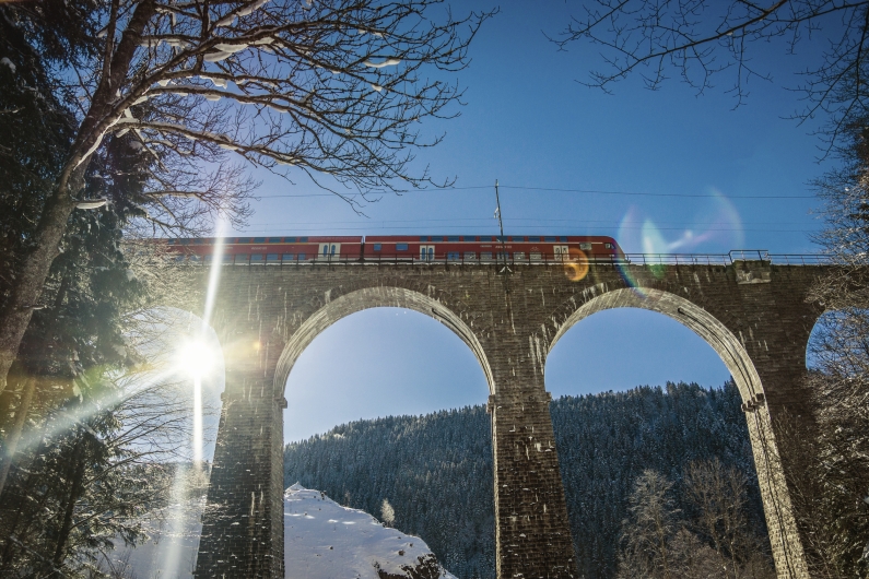 Eine Untersichtaufnahme des Ravennaviadukts im winterlichen Schwarzwald. Zwei massive Steinbögen des Viadukts dominieren das Bild, über die gerade ein roter Regionalzug (Doppelstockwagen) der Höllentalbahn fährt. Die strahlende Sonne steht tief hinter einem Pfeiler auf der linken Seite und erzeugt einen markanten Lichtstern sowie Lens-Flare-Effekte. Der Hang unter der Brücke ist schneebedeckt, und kahle Baumzweige rahmen das Motiv am oberen und linken Bildrand ein. Der Himmel ist wolkenlos und tiefblau.