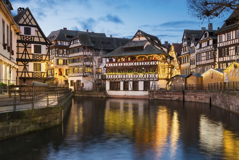 Eine abendliche Aufnahme des historischen Viertels „Petite France“ in Straßburg. Mehrere gut erhaltene Fachwerkhäuser mit ihren charakteristischen dunklen Holzbalken und weißen Fassaden stehen direkt am Ufer des Flusses Ill. Die Gebäude sind warm beleuchtet und teilweise mit weihnachtlicher Dekoration und Lichterketten geschmückt. Im Vordergrund spiegelt sich die goldene Beleuchtung der Fenster und Fassaden auf der ruhigen Wasseroberfläche. Der Himmel zeigt das tiefe Blau der späten Dämmerung mit einigen weichen Wolken.
