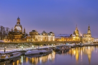 Eine weitwinklige Nachtaufnahme der Dresdner Skyline vom gegenüberliegenden Elbufer aus. Das Stadtpanorama zeigt markante Bauwerke wie die hell erleuchtete Frauenkirche, die Kunstakademie mit ihrer Glaskuppel und die Brühlsche Terrasse. Im Vordergrund liegen mehrere Fahrgastschiffe der Weißen Flotte am verschneiten Terrassenufer vor Anker. Die warmen Lichter der Gebäude spiegeln sich klar auf der Wasseroberfläche der Elbe. Der Himmel ist in das tiefe Blau der späten Dämmerung getaucht.
