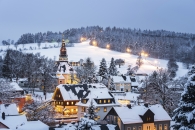 Eine abendliche Panoramaaufnahme des Kurortes Seiffen im Erzgebirge unter einer geschlossenen Schneedecke. Im Zentrum ragt die markante, hell erleuchtete Seiffener Bergkirche mit ihrem dunklen Turm heraus. Die umliegenden Wohnhäuser und Werkstätten haben schneebedeckte Dächer und strahlen warmes Licht aus den Fenstern aus. Im Hintergrund erstreckt sich ein bewaldeter Hang, auf dem eine beleuchtete Skipiste als helles Band durch die verschneiten Bäume verläuft. Der Himmel ist in einem kühlen, dämmerigen Blaugrau gehalten.