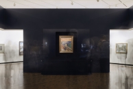 Eine Innenansicht eines modern gestalteten Ausstellungsraums im MUNCH Museum. Im Zentrum einer breiten, dunkelblauen Wand hängt in einer beleuchteten Vitrine das berühmte Gemälde „Der Schrei“ von Edvard Munch. Das Bild zeigt eine Figur mit gequältem Gesichtsausdruck vor einem orange-roten Himmel. Flankiert wird die zentrale Wand von zwei hellen Durchgängen zu weiteren Galerieräumen, in denen links und rechts weitere gerahmte Kunstwerke erkennbar sind. Im Vordergrund sind schemenhaft die Silhouetten mehrerer Museumsbesucher als Bewegungsunschärfe zu sehen.