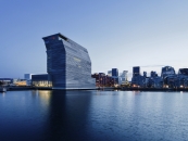 Eine abendliche Weitwinkelaufnahme des neuen Munch-Museums in Oslo. Das markante, 60 Meter hohe Gebäude mit seiner Fassade aus recyceltem, perforiertem Aluminium dominiert die linke Bildhälfte. Der obere Teil des Turms ist charakteristisch zur Seite geneigt. Das Museum steht direkt am Wasser des Oslofjords, in dem sich die Lichter der Stadt spiegeln. Im Hintergrund rechts ist die beleuchtete Skyline des modernen Bjørvika-Viertels mit weiteren zeitgenössischen Hochhäusern zu sehen. Der Himmel ist in ein klares, tiefes Blau der Dämmerung getaucht.