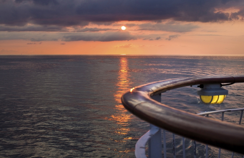 Eine maritime Aufnahme von einem Schiffsdeck aus bei Sonnenuntergang. Im rechten Vordergrund ist die hölzerne Reling des Decks zu sehen, an der eine leuchtende, gelbe Außenlampe angebracht ist. Die Sonne steht tief am Horizont hinter einer dunklen Wolkenschicht und wirft eine glitzernde Lichtstraße auf die leicht bewegte Wasseroberfläche. Der Himmel ist in warme Orange- und Gelbtöne getaucht, während das Meer und die Wolken in dunklen Blau- und Grautönen erscheinen.