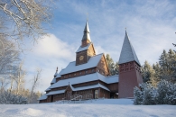 Die verschneite Gustav-Adolf-Stabkirche in Goslar-Hahnenklee bei sonnigem Winterwetter. Das archaische Kirchengebäude aus dunklem Fichtenholz ist nach norwegischem Vorbild erbaut und verfügt über mehrere gestaffelte Dachebenen sowie einen freistehenden Glockenturm auf der rechten Seite.