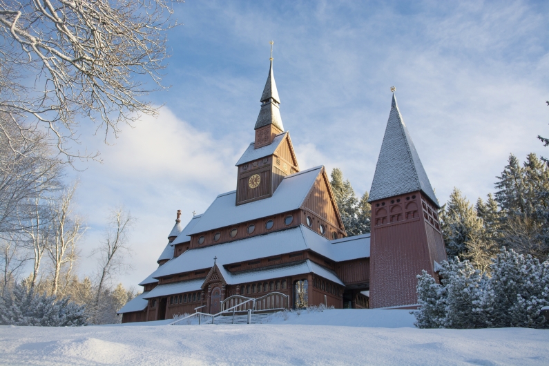 Die verschneite Gustav-Adolf-Stabkirche in Goslar-Hahnenklee bei sonnigem Winterwetter. Das archaische Kirchengebäude aus dunklem Fichtenholz ist nach norwegischem Vorbild erbaut und verfügt über mehrere gestaffelte Dachebenen sowie einen freistehenden Glockenturm auf der rechten Seite.