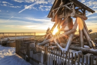 Die hölzerne Friedensglocke auf dem Fichtelberg (Sachsen) in einer verschneiten Winterlandschaft bei Sonnenuntergang. Die tiefstehende Sonne bricht als strahlender Stern durch das Gebälk des Glockenstuhls und taucht die Szenerie in warmes, goldenes Licht. Im Hintergrund ist eine Aussichtsplattform mit Metallgeländer unter einem blau-weiß bewölkten Himmel zu sehen.