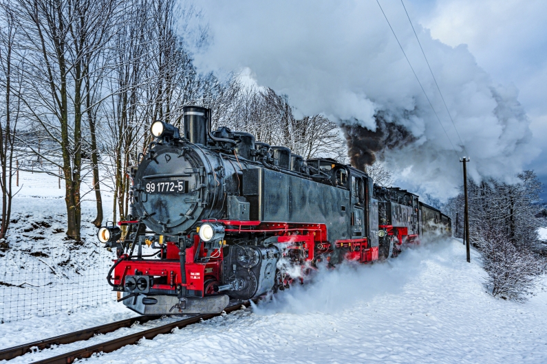 Eine historische Dampflokomotive der Baureihe 99 (Nummer 99 1772-5) der Fichtelbergbahn fährt mit einer großen, weißen Dampfwolke durch eine tief verschneite Winterlandschaft im Erzgebirge. Die schwarz-rote Lokomotive ist von vorne rechts zu sehen, wie sie auf den Schienen durch den hohen Schnee dampft, während im Hintergrund kahle, schneebedeckte Bäume unter einem grauen Winterhimmel stehen.