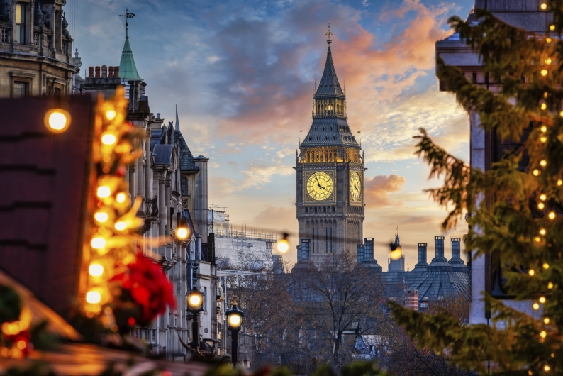Blick auf den beleuchteten Elizabeth Tower (Big Ben) in London während der Abenddämmerung. Im Vordergrund sind unscharf fokussierte Weihnachtsdekorationen mit Lichterketten und Tannenzweigen zu sehen, die eine festliche Atmosphäre schaffen. Der Himmel im Hintergrund leuchtet in Blau- und Rosatönen, während die Zifferblätter der Uhr hell erstrahlen.