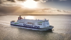 Eine große, weiß-blaue Stena Line Kombifähre fährt bei Gegenlicht auf dem offenen Meer. Das Schiff ist von der Steuerbordseite zu sehen, mit dem markanten roten Schornstein und dem Schriftzug „Stena Line“ am Rumpf. Die tiefstehende Sonne spiegelt sich glitzernd auf der Wasseroberfläche und bricht durch eine leichte Wolkendecke am Horizont.