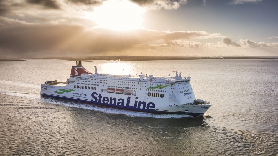 Eine große, weiß-blaue Stena Line Kombifähre fährt bei Gegenlicht auf dem offenen Meer. Das Schiff ist von der Steuerbordseite zu sehen, mit dem markanten roten Schornstein und dem Schriftzug „Stena Line“ am Rumpf. Die tiefstehende Sonne spiegelt sich glitzernd auf der Wasseroberfläche und bricht durch eine leichte Wolkendecke am Horizont.
