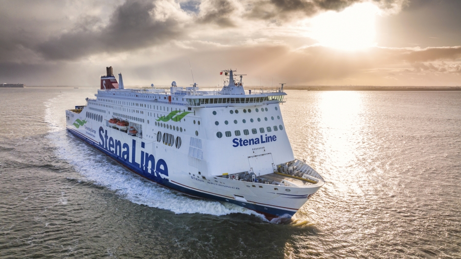 Eine große, weiß-blaue Stena Line Kombifähre fährt bei Gegenlicht auf dem offenen Meer. Das Schiff ist von der Steuerbordseite zu sehen, mit dem markanten roten Schornstein und dem Schriftzug „Stena Line“ am Rumpf. Die tiefstehende Sonne spiegelt sich glitzernd auf der Wasseroberfläche und bricht durch eine leichte Wolkendecke am Horizont.