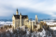 Das verschneite Schloss Neuschwanstein in den bayerischen Alpen bei klarem Winterwetter. Die hellen Kalksteinfassaden und die markanten blauen Türme des Märchenschlosses heben sich kontrastreich von den schneebedeckten Felsen und dunklen Tannen im Vordergrund ab. Im Hintergrund erstreckt sich eine weite, winterliche Hügellandschaft unter einem blau-weiß bewölkten Himmel.