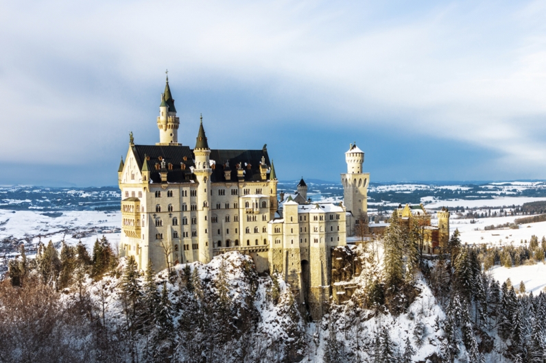 Das verschneite Schloss Neuschwanstein in den bayerischen Alpen bei klarem Winterwetter. Die hellen Kalksteinfassaden und die markanten blauen Türme des Märchenschlosses heben sich kontrastreich von den schneebedeckten Felsen und dunklen Tannen im Vordergrund ab. Im Hintergrund erstreckt sich eine weite, winterliche Hügellandschaft unter einem blau-weiß bewölkten Himmel.