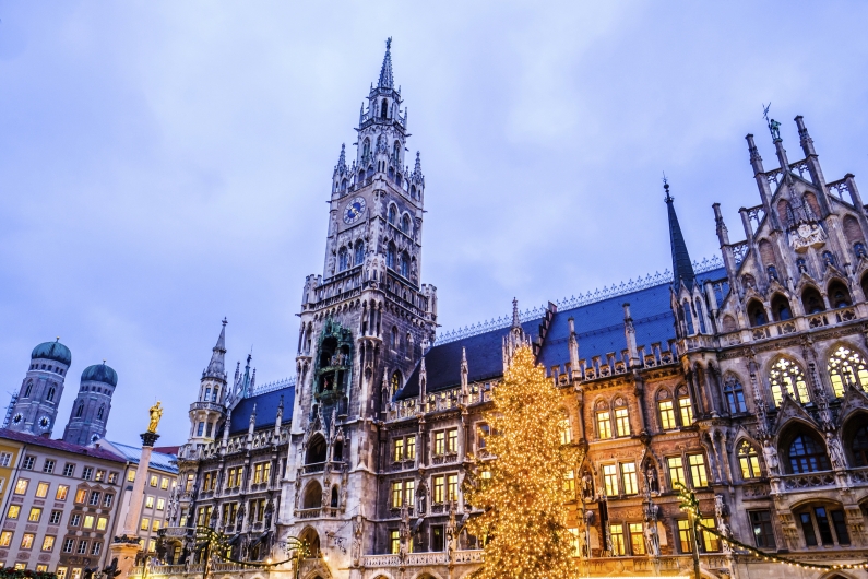 Das festlich beleuchtete Neue Rathaus am Münchener Marienplatz zur Zeit des Christkindlmarktes in der Abenddämmerung. Im Vordergrund erstrahlt ein großer, mit unzähligen Lichtern geschmückter Weihnachtsbaum. Links im Hintergrund ragen die charakteristischen Zwiebeltürme der Frauenkirche in den dunkelblauen Abendhimmel.
