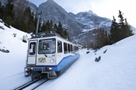 Ein blau-weißer Triebwagen der Bayerischen Zugspitzbahn (Nummer 14) fährt auf einer verschneiten Gleisanlage bergab. Die Bahn trägt das bayerische Staatswappen auf der Frontseite. Im Hintergrund ragen die massiven, schneebedeckten Felswände des Wettersteingebirges steil unter einem hellen Himmel auf, flankiert von schneebedeckten Nadelbäumen.
