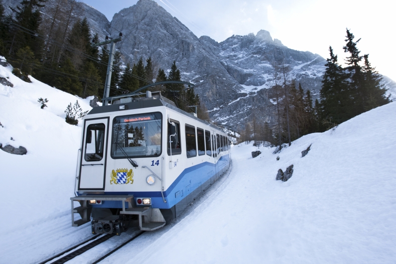 Ein blau-weißer Triebwagen der Bayerischen Zugspitzbahn (Nummer 14) fährt auf einer verschneiten Gleisanlage bergab. Die Bahn trägt das bayerische Staatswappen auf der Frontseite. Im Hintergrund ragen die massiven, schneebedeckten Felswände des Wettersteingebirges steil unter einem hellen Himmel auf, flankiert von schneebedeckten Nadelbäumen.