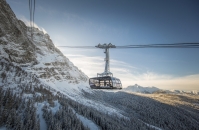 Eine moderne, vollverglaste Kabine der Seilbahn Zugspitze schwebt an Tragseilen vor der schneebedeckten Nordflanke des Berges. Im Hintergrund erstreckt sich ein weites Panorama der winterlichen Alpen mit verschneiten Nadelwäldern im Tal unter einem klaren, sonnigen Himmel. Die Kabine trägt die Aufschrift „Zugspitze“.