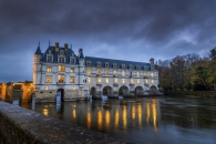 Das beleuchtete Schloss Chenonceau in Frankreich während der blauen Stunde. Das Schloss ist auf massiven Bögen direkt über den Fluss Cher gebaut, dessen ruhige Wasseroberfläche die hell erleuchteten Fenster spiegelt. Ein bewölkter, dunkler Abendhimmel und herbstliche Bäume im Hintergrund umrahmen die historische Architektur.