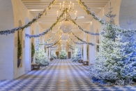 Die festlich für Weihnachten geschmückte Große Galerie von Schloss Chenonceau. Der lange Saal mit seinem charakteristischen schwarz-weißen Fliesenboden ist gesäumt von künstlich beschneiten Tannenbäumen. Von der Balkendecke hängen goldene Kronleuchter, große weiße Lichtkugeln und girlandenartig gespannte Tannen- und Blumenzweige, die eine tunnelartige, winterliche Atmosphäre schaffen.