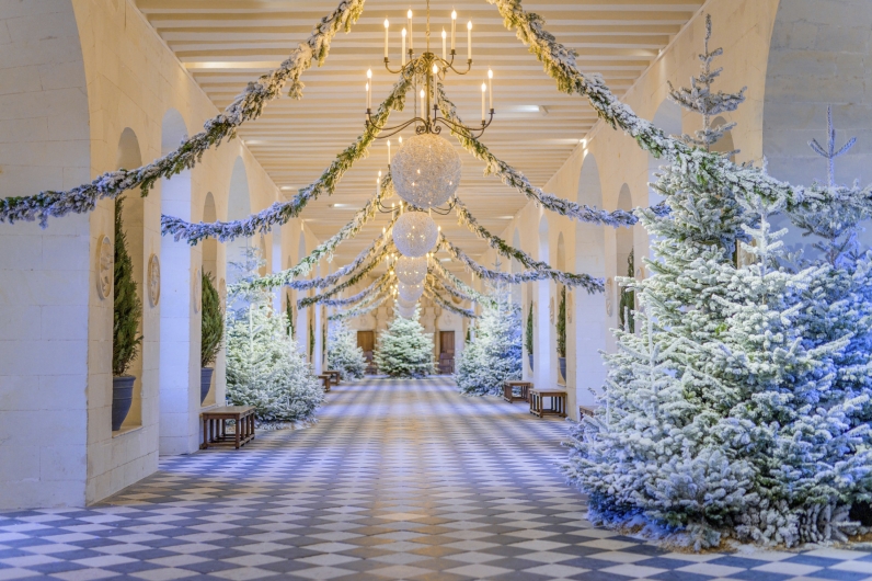 Die festlich für Weihnachten geschmückte Große Galerie von Schloss Chenonceau. Der lange Saal mit seinem charakteristischen schwarz-weißen Fliesenboden ist gesäumt von künstlich beschneiten Tannenbäumen. Von der Balkendecke hängen goldene Kronleuchter, große weiße Lichtkugeln und girlandenartig gespannte Tannen- und Blumenzweige, die eine tunnelartige, winterliche Atmosphäre schaffen.