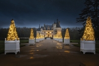 Blick auf das beleuchtete Schloss Chenonceau bei Nacht. Ein langer Weg führt direkt auf das Schloss zu, gesäumt von zwei Reihen kegelförmig geschnittener Lichterbäume in weißen Kübeln, die warmes, goldenes Licht ausstrahlen. Der dunkle Nachthimmel darüber ist sternenklar, und die herbstlichen Bäume des Schlossparks umrahmen die Szenerie.