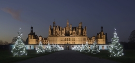 Frontalansicht des hell erleuchteten Schlosses Chambord bei Nacht. Ein zentraler Weg führt auf das monumentale Renaissance-Bauwerk zu, flankiert von zwei Reihen prächtig mit weißen Lichterketten geschmückter Tannenbäume. Das Schloss besticht durch seine markante Dachlandschaft mit zahlreichen Türmen, Schornsteinen und Laternen unter einem tiefblauen Nachthimmel.