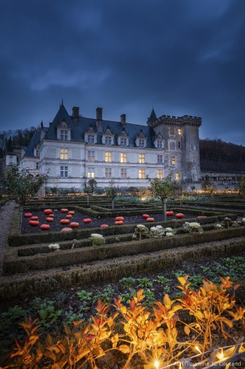 Blick über einen terrassierten Renaissancegarten auf das hell erleuchtete Schloss Villandry in der Abenddämmerung. Im Vordergrund sind rechtwinklige Gartenbeete zu sehen, die mit niedrigen Hecken eingefasst sind; in einem Beet liegen mehrere leuchtend orangefarbene Kürbisse. Das Schloss im Hintergrund zeigt eine helle Fassade mit vielen beleuchteten Fenstern, steilen Schieferdächern und einem markanten Wehrturm auf der rechten Seite unter einem dunkelblauen Wolkenhimmel.