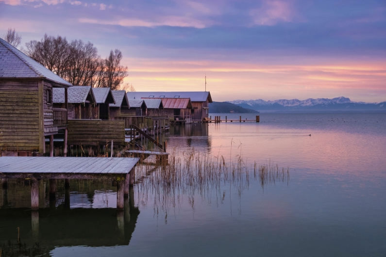 Eine winterliche Weitwinkelaufnahme eines Sees in Oberbayern (vermutlich am Starnberger See oder Ammersee) während der Dämmerung. Im linken Vordergrund stehen mehrere rustikale, hölzerne Bootshäuser auf Stelzen über dem Wasser, deren Dächer mit einer leichten Schneeschicht bedeckt sind. Von den Hütten führen schmale Holzstege in den See. Im ruhigen Wasser spiegeln sich die Farben des Himmels und einzelne Halme von vertrocknetem Schilf ragen aus der Wasseroberfläche empor. Im Hintergrund erstreckt sich am Horizont das Panorama der schneebedeckten Alpen unter einem dramatischen Abendhimmel, der in sanften Violett-, Rosa- und Blautönen leuchtet. Die gesamte Szenerie wirkt friedlich und eiskalt.