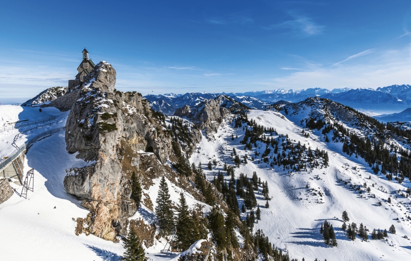 Eine alpine Panoramaaufnahme vom Gipfel des Wendelsteins im Winter bei strahlendem Sonnenschein und tiefblauem Himmel. Auf der Spitze eines markanten, grauen Felsturms im linken Bilddrittel thront die kleine Wendelsteinkirche. Die steilen Felshänge sind teilweise schneebedeckt. Nach rechts und in die Tiefe erstreckt sich eine weite, verschneite Berglandschaft mit Skipisten und dunklen Nadelbäumen. Am Horizont ist die Kette der schneebedeckten Zentralalpen unter einem klaren Himmel zu sehen. Im Vordergrund links erkennt man einen Teil der befestigten Aussichtsplattform mit Geländer.