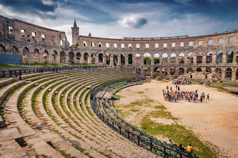 Eine Weitwinkelaufnahme des Innenraums des Amphitheaters von Pula in Kroatien bei Tageslicht. Das Bild zeigt die gut erhaltenen, halbkreisförmigen Steinstufen der Zuschauerränge im linken Vordergrund, die zur sandigen Arena in der Mitte abfallen. Dort steht eine Gruppe von Touristen. Den Hintergrund bildet die imposante, mehrstöckige Außenmauer aus hellem Kalkstein mit ihren zahlreichen Rundbogenöffnungen. Über die Mauer ragt links im Hintergrund die Spitze eines Kirchturms hinaus. Der Himmel ist blau mit einigen weißen und grauen Wolken formationen.