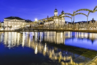 Weitwinkelaufnahme des beleuchteten Rathausstegs in Luzern zur blauen Stunde. Die Fußgängerbrücke überspannt im Vordergrund die dunkle Reuss, in deren Wasser sich die Lichter der Stadt golden spiegeln. Über der Brücke wölben sich mehrere mit warmweißen Lichterketten geschmückte Girlandenbögen. Im Hintergrund ragen die markanten Zwiebeltürme der barocken Jesuitenkirche hell erleuchtet in den tiefblauen Abendhimmel. Links am Ufer ist die strahlend weiße Fassade des Luzerner Theaters zu sehen.