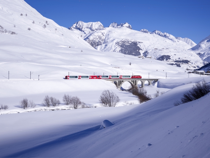 Eine Panoramaaufnahme des rot-weißen Glacier Express in einer tief verschneiten Hochgebirgslandschaft der Schweizer Alpen bei strahlend blauem Himmel. Der Zug, bestehend aus einer roten Elektrolokomotive und mehreren modernen Panoramawagen mit großen Glasfronten, überquert gerade eine steinerne Bogenbrücke. Die gesamte Umgebung ist von einer dicken, unberührten Schneeschicht bedeckt, die in der Sonne glänzt und sanfte Schatten wirft. Im Hintergrund ragen imposante, schroffe Gebirgsmassive mit schneebedeckten Gipfeln und steilen Felsflanken auf. Vereinzelt sind im Tal unterhalb der Brücke schneefreie, dunkle Sträucher und Bäume zu erkennen.