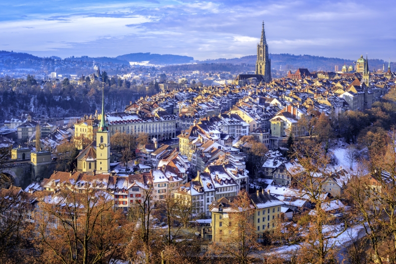 Weitläufige Panoramaaufnahme der Berner Altstadt im Winter aus erhöhter Perspektive. Die dichte historische Bebauung mit ihren charakteristischen ziegelroten Dächern ist leicht mit Schnee bestäubt. Markante architektonische Wahrzeichen wie das Berner Münster mit seinem hohen, gotischen Turm und die grüne Kuppel des Bundeshauses ragen aus dem Häusermeer hervor. Im Vordergrund sind herbstlich braune, teils schneebedeckte Baumwipfel zu sehen, die den Blick auf das Stadtbild einrahmen. Die Szenerie liegt in sanftem Tageslicht unter einem blauen, leicht bewölkten Himmel, während im Hintergrund bewaldete Hügelketten das Stadtgebiet begrenzen.