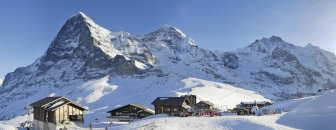 Weitwinkel-Panoramaaufnahme einer alpinen Winterlandschaft im Berner Oberland. Im Vordergrund befindet sich die verschneite Station Kleine Scheidegg mit mehreren rustikalen Holzhütten, Chalets und einer Bergbahnstation. Vor den Gebäuden sind vereinzelt bunte Skier und Ausrüstungsgegenstände im Schnee erkennbar. Majestätisch ragen im Hintergrund die drei berühmten, schneebedeckten Gipfel der Schweizer Alpen auf: links die markante Nordwand des Eigers, daneben der Mönch und die Jungfrau. Die felsigen Steilwände kontrastieren scharf mit den weißen Schneefeldern unter einem klaren, hellblauen Himmel.