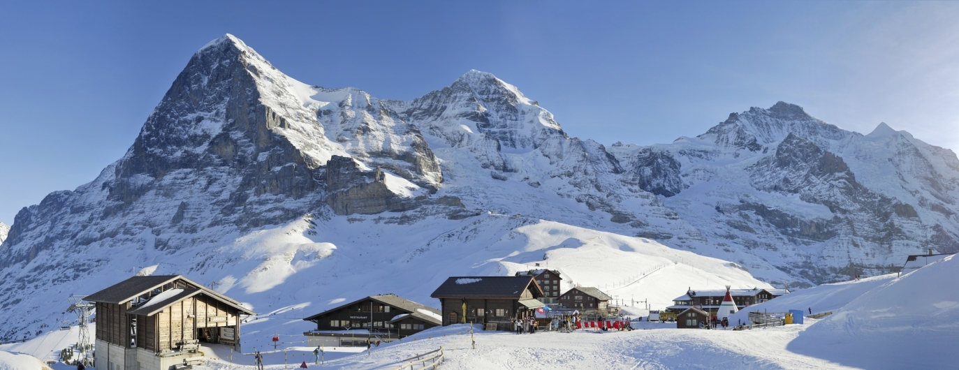 Weitwinkel-Panoramaaufnahme einer alpinen Winterlandschaft im Berner Oberland. Im Vordergrund befindet sich die verschneite Station Kleine Scheidegg mit mehreren rustikalen Holzhütten, Chalets und einer Bergbahnstation. Vor den Gebäuden sind vereinzelt bunte Skier und Ausrüstungsgegenstände im Schnee erkennbar. Majestätisch ragen im Hintergrund die drei berühmten, schneebedeckten Gipfel der Schweizer Alpen auf: links die markante Nordwand des Eigers, daneben der Mönch und die Jungfrau. Die felsigen Steilwände kontrastieren scharf mit den weißen Schneefeldern unter einem klaren, hellblauen Himmel.