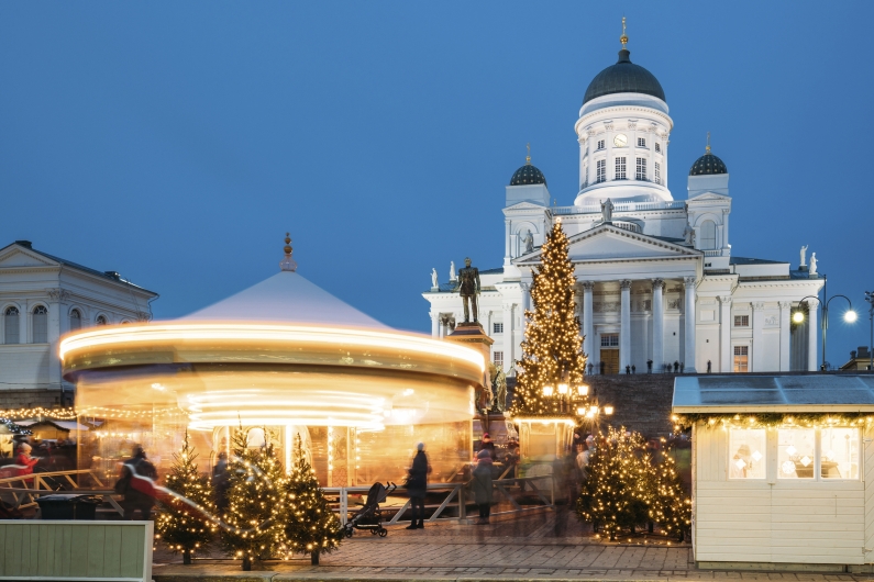 Abendliche Langzeitbelichtung des Weihnachtsmarktes in Helsinki während der blauen Stunde. Im Zentrum des Vordergrunds ist ein hell erleuchtetes Karussell in Bewegung zu sehen, dessen Lichter durch die Belichtungszeit als leuchtende Streifen erscheinen. Daneben steht ein großer, prachtvoll mit warmweißen Lichtern geschmückter Tannenbaum. Im Hintergrund thront der majestätische, weiß leuchtende Dom von Helsinki mit seinen markanten grünen Kuppeln unter einem tiefblauen Abendhimmel. Der Platz ist belebt mit Passanten, die zwischen kleinen, hellen Marktbuden und festlich beleuchteten Weihnachtsbäumen flanieren.