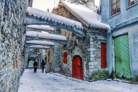 Blick in die schmale, mittelalterliche St.-Katharinen-Passage (Katariina käik) in Tallinn im Winter. Charakteristische, massive Steinbögen überspannen die Gasse und sind, ebenso wie die Dächer, mit einer dicken Schneeschicht bedeckt. Die historischen Fassaden aus Bruchstein und farbigem Putz – darunter eine auffällige grüne und rote Holztür – säumen den schneebedeckten Weg. In der Ferne spazieren zwei Personen unter den Bögen hindurch. Die Szenerie wirkt durch die Kombination aus altem Mauerwerk und frischem Schnee besonders verwunschen und authentisch.
