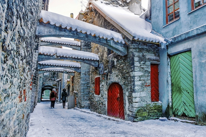 Blick in die schmale, mittelalterliche St.-Katharinen-Passage (Katariina käik) in Tallinn im Winter. Charakteristische, massive Steinbögen überspannen die Gasse und sind, ebenso wie die Dächer, mit einer dicken Schneeschicht bedeckt. Die historischen Fassaden aus Bruchstein und farbigem Putz – darunter eine auffällige grüne und rote Holztür – säumen den schneebedeckten Weg. In der Ferne spazieren zwei Personen unter den Bögen hindurch. Die Szenerie wirkt durch die Kombination aus altem Mauerwerk und frischem Schnee besonders verwunschen und authentisch.