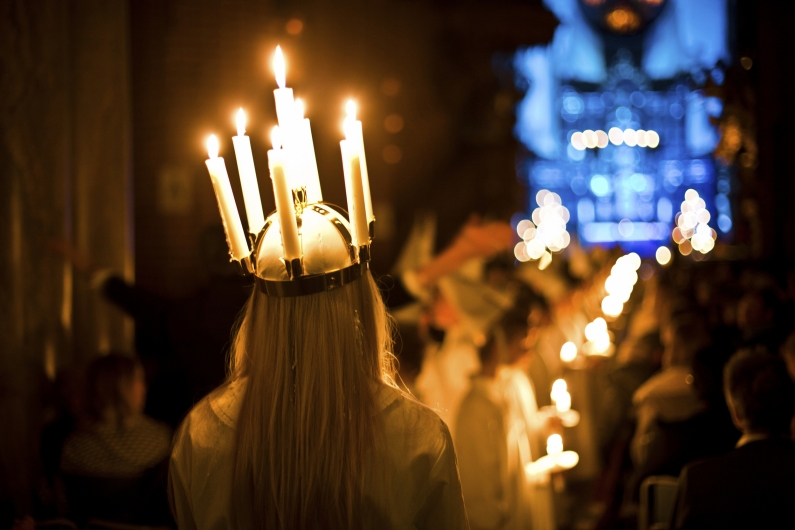 Nahaufnahme von hinten auf eine junge Frau mit langem hellem Haar, die während einer Lucia-Prozession eine brennende Kerzenkrone trägt. Die sechs weißen Kerzen werfen einen warmen, goldenen Schein auf ihren Kopf und ihre weißen Schultern. Im unscharfen Hintergrund sind weitere Teilnehmer in weißen Gewändern erkennbar, die brennende Kerzen in den Händen halten und in einer Reihe durch den dunklen Kirchenraum ziehen. Ein blau beleuchteter Altarbereich im Hintergrund bildet einen kühlen Kontrast zu den vielen warmen Lichtpunkten der Kerzen, was eine feierliche und andächtige Atmosphäre schafft.