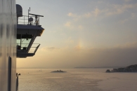 Seitenansicht der Brückennock einer großen Fähre, die links ins Bild ragt. Man erkennt die Reling, Antennen und Radarsysteme des Schiffes vor einem weiten, morgendlichen Himmel. Der Blick schweift über eine weite, teils zugefrorene Wasserfläche, aus der vereinzelte kleine Schärinseln mit dunklen Bäumen herausragen. Die tiefstehende Sonne taucht die Szenerie in ein warmes, gelblich-rosa Licht, das sich sanft auf der Eisoberfläche und den leichten Wolken spiegelt. In der Bildmitte ist ein schwacher, vertikaler Lichtreflex am Himmel zu sehen.