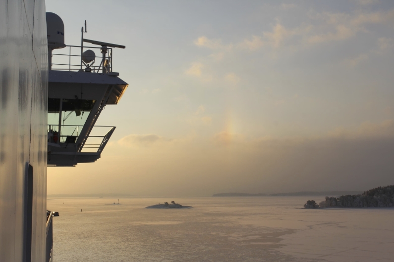 Seitenansicht der Brückennock einer großen Fähre, die links ins Bild ragt. Man erkennt die Reling, Antennen und Radarsysteme des Schiffes vor einem weiten, morgendlichen Himmel. Der Blick schweift über eine weite, teils zugefrorene Wasserfläche, aus der vereinzelte kleine Schärinseln mit dunklen Bäumen herausragen. Die tiefstehende Sonne taucht die Szenerie in ein warmes, gelblich-rosa Licht, das sich sanft auf der Eisoberfläche und den leichten Wolken spiegelt. In der Bildmitte ist ein schwacher, vertikaler Lichtreflex am Himmel zu sehen.