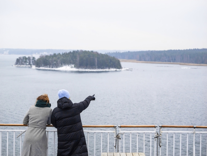 Rückansicht von zwei Personen, die an der Reling eines Schiffsdecks stehen und auf das Meer blicken. Eine Person in einer schwarzen Steppjacke und hellblauer Mütze deutet mit ausgestrecktem Arm auf eine kleine, bewaldete Insel im Mittelgrund. Die zweite Person trägt einen grauen Mantel und einen dunkelgrünen Schal. Die Insel zeigt dunkle Nadelbäume und Reste von Schnee am Ufer. Im Hintergrund erstreckt sich eine weitere, flache Küstenlinie unter einem wolkenverhangenen, hellgrauen Himmel. Das Wasser ist ruhig und dunkelblau gefärbt.