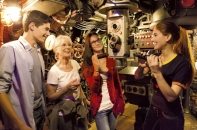 Innenaufnahme aus der engen, technisch detailreichen Kulisse des U-Boots aus dem Film „Das Boot“. Eine Museumsführerin auf der rechten Seite erklärt einer dreiköpfigen Besuchergruppe die Armaturen. Die Führerin ist eine junge Frau mit einem Zopf, die ein dunkles T-Shirt mit gelbem Saum trägt und ein zylindrisches Bauteil in den Händen hält. Die Besucher – ein Jugendlicher im hellblauen Hemd, eine ältere Frau mit Brille im weißen T-Shirt und eine junge Frau mit Brille im roten Cardigan – blicken interessiert und lächelnd zu ihr. Die Umgebung ist dicht gedrängt mit historischen technischen Geräten, darunter ein Periskop in der Bildmitte, Rohrleitungen, Ventile, Kabelstränge an der Decke und beleuchtete Anzeigeinstrumente. Die Szene ist in warmes, atmosphärisches Licht getaucht.