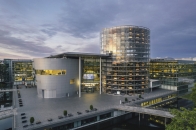 Architekturfotografie aus erhöhter Perspektive bei bewölktem Abendhimmel. Zu sehen ist der moderne Gebäudekomplex der Gläsernen Manufaktur. Markantestes Element auf der rechten Seite ist ein hoher, vollkommen transparenter Zylinderturm aus Glas, in dessen Inneren zahlreiche Autos auf mehreren Etagen übereinander eingeparkt und beleuchtet sind. Links daneben schließt sich der Haupteingang mit einer geschwungenen, silber-grauen Fassade an. Über dem Eingangsbereich leuchtet der Schriftzug „DIE GLÄSERNE MANUFAKTUR Home of ID.“. Vor dem Gebäude erstreckt sich ein großer, leerer Vorplatz aus grauen Steinplatten, der von Wasserflächen und Gehwegen mit Geländern umgeben ist. Im Inneren der gläsernen Fabrikgebäude brennt helles Licht.