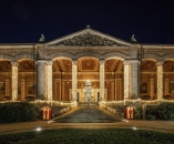 Symmetrische Nachtaufnahme der historischen Trinkhalle in Baden-Baden im weihnachtlichen Festgewand. Die monumentale, klassizistische Fassade wird von einer offenen Vorhalle mit einer Reihe mächtiger, korinthischer Säulen geprägt. Jede dieser Säulen ist vollständig spiralartig mit warmweiß leuchtenden Lichterketten umwickelt. Eine breite, halbrunde Freitreppe aus Stein führt zum zentralen Eingang empor. Auf dem Treppenabsatz steht ein großer, dicht geschmückter und hell strahlender Weihnachtsbaum hinter einer kleinen Holzbarriere. Auch die Balustraden links und rechts der Treppe sind mit Lichterketten geschmückt. Am Fuß der Treppe steht links und rechts jeweils ein dekoratives, rot-weißes Häuschen mit einer Nussknacker-Figur darin. Das Giebeldreieck (Timpanon) über dem Eingang zeigt ein historisches Steinrelief. Der Nachthimmel darüber ist tiefschwarz und klar. Im Vordergrund befindet sich eine dunkle Rasenfläche.