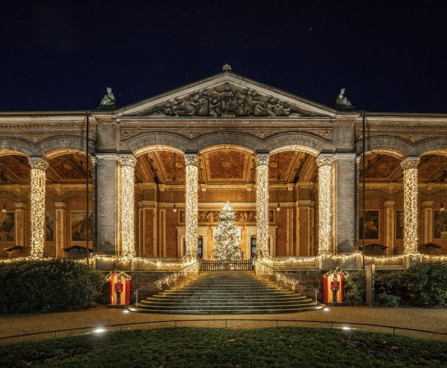 Symmetrische Nachtaufnahme der historischen Trinkhalle in Baden-Baden im weihnachtlichen Festgewand. Die monumentale, klassizistische Fassade wird von einer offenen Vorhalle mit einer Reihe mächtiger, korinthischer Säulen geprägt. Jede dieser Säulen ist vollständig spiralartig mit warmweiß leuchtenden Lichterketten umwickelt. Eine breite, halbrunde Freitreppe aus Stein führt zum zentralen Eingang empor. Auf dem Treppenabsatz steht ein großer, dicht geschmückter und hell strahlender Weihnachtsbaum hinter einer kleinen Holzbarriere. Auch die Balustraden links und rechts der Treppe sind mit Lichterketten geschmückt. Am Fuß der Treppe steht links und rechts jeweils ein dekoratives, rot-weißes Häuschen mit einer Nussknacker-Figur darin. Das Giebeldreieck (Timpanon) über dem Eingang zeigt ein historisches Steinrelief. Der Nachthimmel darüber ist tiefschwarz und klar. Im Vordergrund befindet sich eine dunkle Rasenfläche.